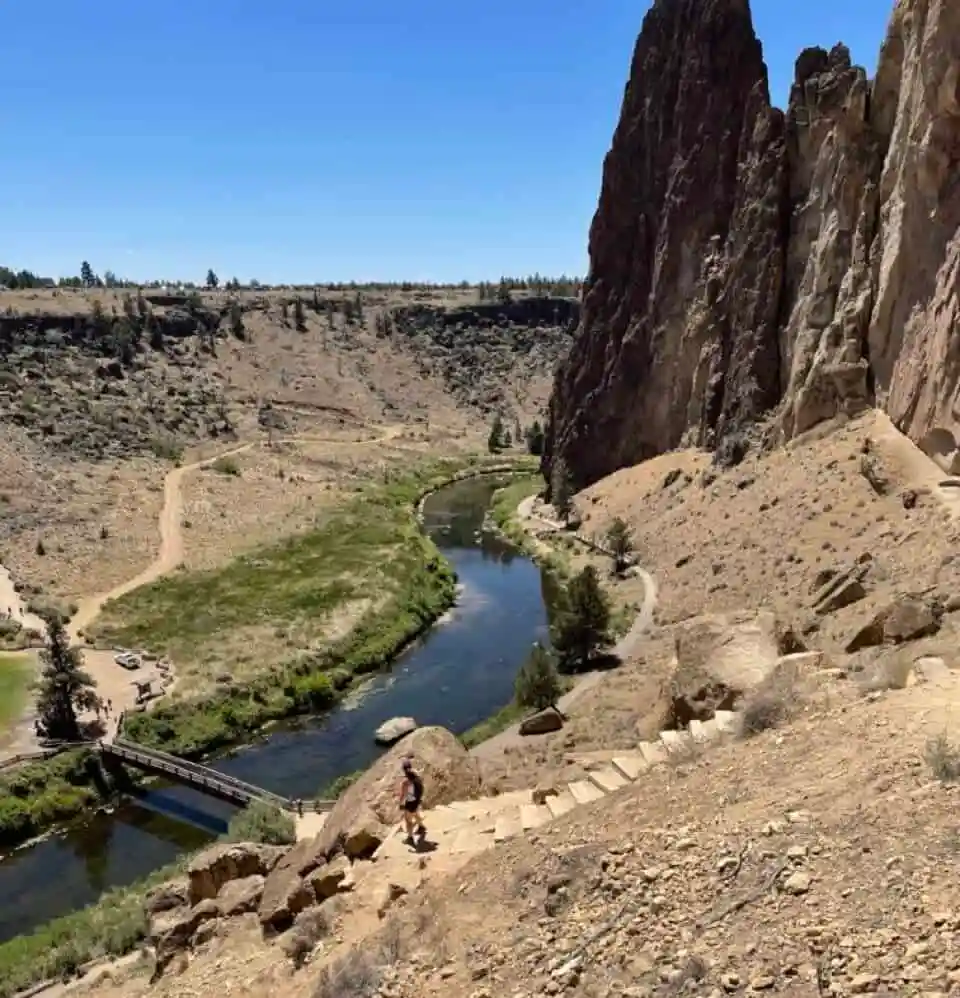 A person climbs stone steps along a dry hillside under a clear blue sky—a classic scene from hikes near Bend Oregon. Below, a winding river flows past green grass, while tall, jagged rock formations and walking paths define the stunning landscape.