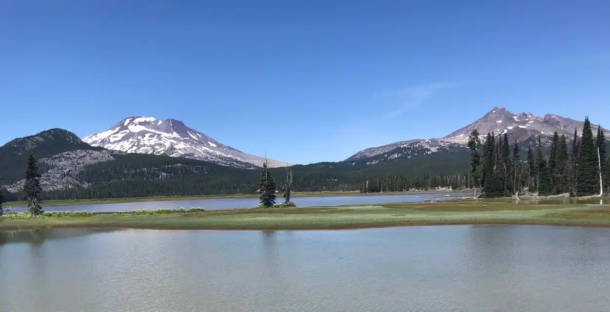 A calm lake in the foreground reflects a clear blue sky, with patches of green grass and scattered pine trees on the shore. In the background, two snow-patched mountains rise above dense forest—perfect for hikes near Bend Oregon in a serene, natural landscape.