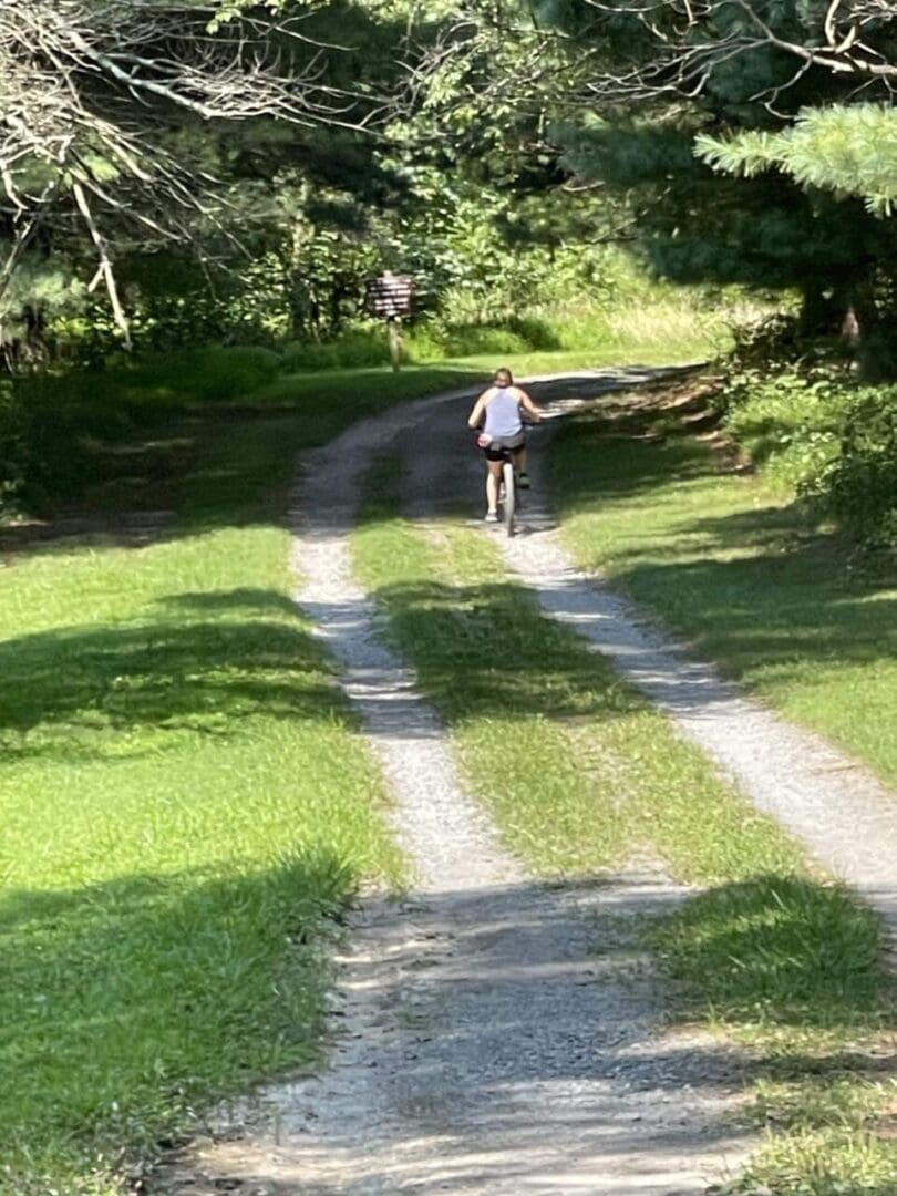 A person wearing a light-colored tank top and dark shorts rides a bicycle along the sunlit Little Bennett Bike Trails, where dappled sunlight filters through tall trees and lush green grass lines the gravel path.