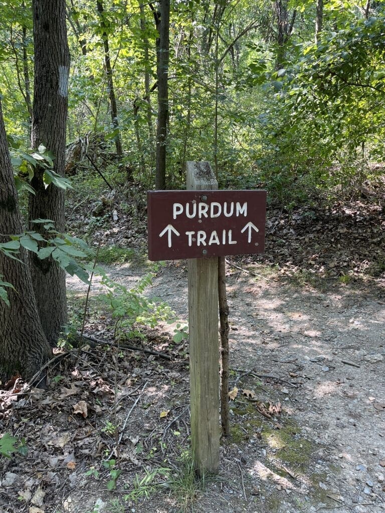 A wooden post holds a brown sign reading PURDUM TRAIL in white letters with two arrows pointing up. Surrounded by trees and foliage, this marker is part of the scenic Little Bennett Bike Trails, with a dirt path winding through the forested background.