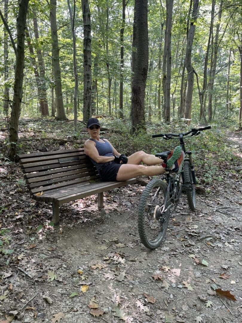 A person in athletic gear reclines on a wooden bench along the Little Bennett Bike Trails, resting their legs on a mountain bike. They wear a cap, sleeveless shirt, shorts, and sneakers beneath tall trees with green foliage and scattered leaves.