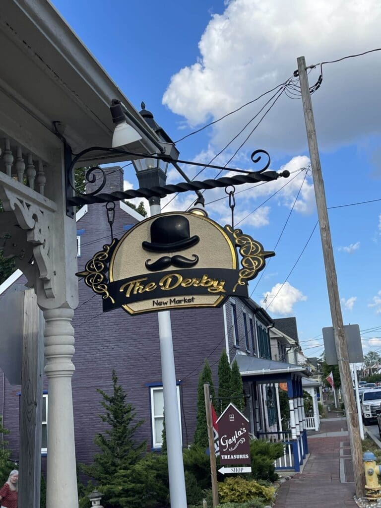 A decorative hanging sign reads “The Derby New Market” with a black bowler hat graphic above the text. The sign is attached to a white post near a brick building, sidewalk, and blue sky—perfect for those exploring tree trekkers Frederick MD.