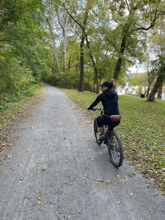 A person wearing a black outfit and cap rides a bicycle on a gravel path through a lush, green forest along one of the scenic bike trails near Frederick MD. Fallen leaves border the path, trees line both sides, and a river is partially visible in the background.