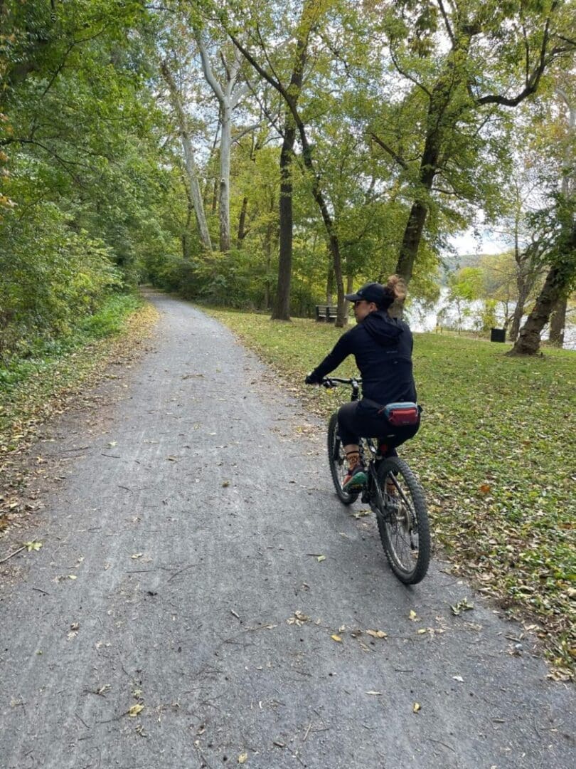 A person wearing a black outfit and cap rides a bicycle on a gravel path through a lush, green forest along one of the scenic bike trails near Frederick MD. Fallen leaves border the path, trees line both sides, and a river is partially visible in the background.