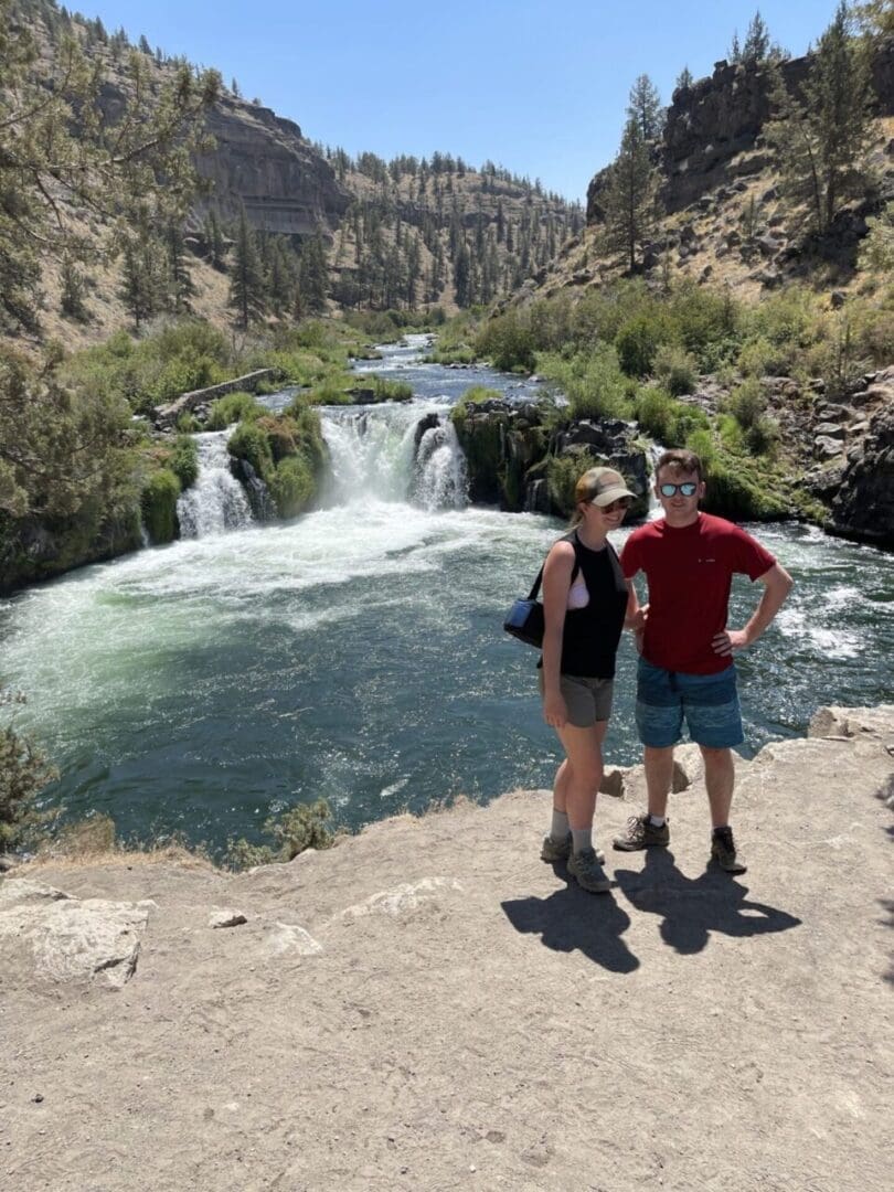 A woman and man stand side by side on a dirt path near a lush river and waterfall, surrounded by green trees and rocky hills. Both wear sunglasses and casual hiking clothes, soaking up the sun on one of many scenic Bend hikes.