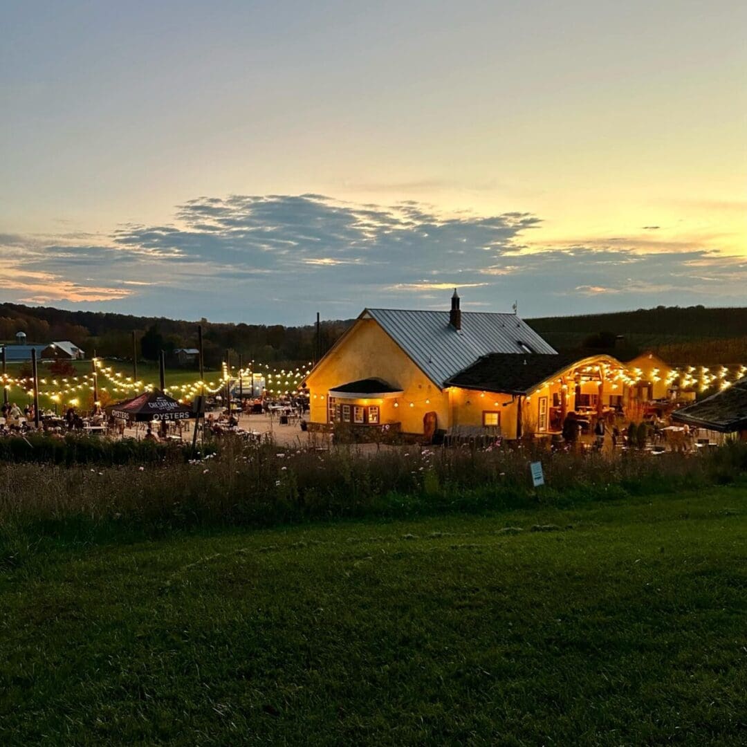 a Yellow Building with String Lights Outside Glows Warmly at Sunset People Gather at Outdoor Tables Enjoying the Cozy Atmosphere Typical of Frederick Md Wineries Rolling Hills and Fields Surround the Scene Beneath a Cloudy Sunlit Sky