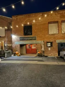 a Brick Bar with String Lights Overhead Features a Sign Reading Mcclintocks Back Bar Above Red Double Doors Pumpkins Sit by the Entrance with a Barrel and Umbrella Nearby Its Nighttime Hinting at the Secret Charm Inside While Cars Line the Sides