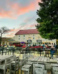 Outdoor dining area with wooden tables and chairs, some occupied by people enjoying meals and conversation. Nearby greenery and a large beige building set the scene beneath a colorful sunset—perfect for relaxing after visiting Frederick MD Wineries.