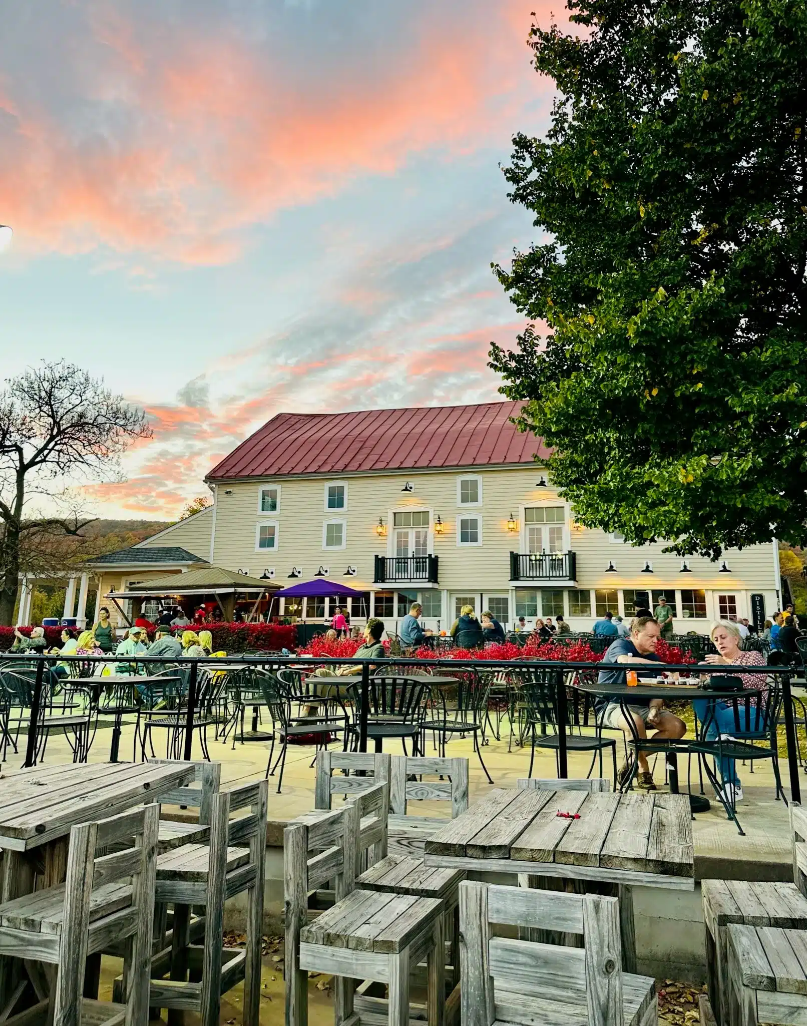 Outdoor dining area with wooden tables and chairs, some occupied by people enjoying meals and conversation. Nearby greenery and a large beige building set the scene beneath a colorful sunset—perfect for relaxing after visiting Frederick MD Wineries.