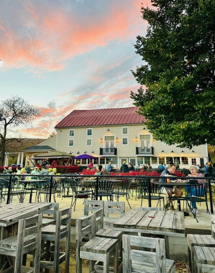 Outdoor Restaurant Scene at Sunset with Wooden Tables and Chairs People Dining and Chatting Near Vibrant Flowerbeds Reminiscent of Frederick Md Wineries a Large Beige Building with a Red Roof in the Background and a Tree with Green Leaves on the Right