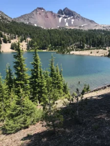 A clear, blue alpine lake is surrounded by green pine trees. Beyond the lake, a rugged mountain with patches of snow rises against a bright blue sky—classic scenery found on the best Bend hikes for those seeking peaceful wilderness.