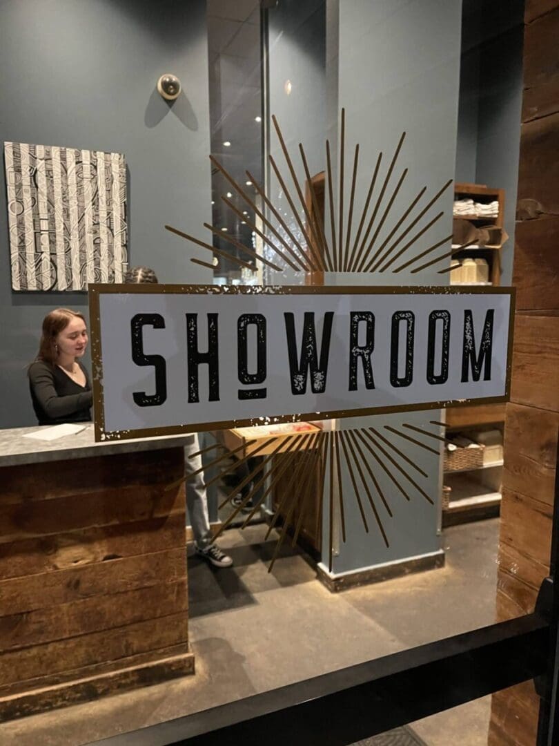 a Glass Door with a Large Showroom Sign and Gold Rays Welcomes You Inside a Woman Stands Behind a Wood Reception Desk Shelves Display Items and the Dark Gray Walls Echo the Stylish Decor Found in Restaurants in Frederick Md