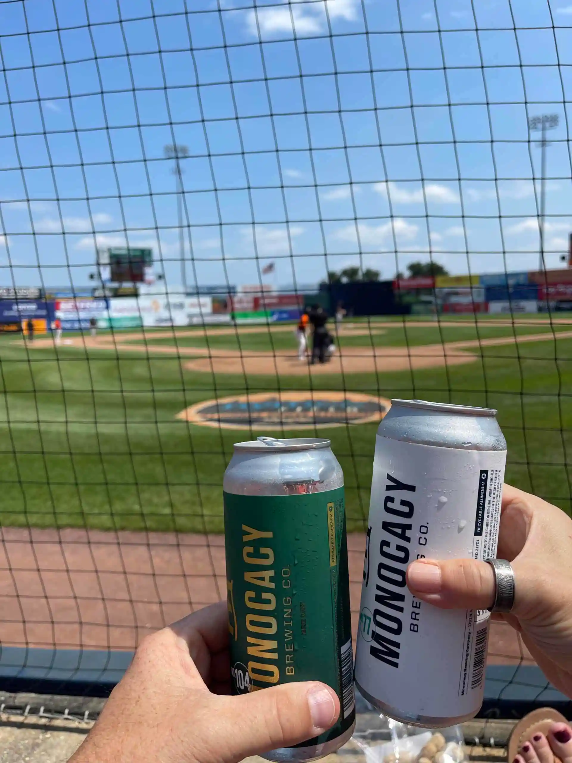 Two hands toast with cans of Monocacy Brewing Co. beer at a baseball stadium, field and players in view under a blue sky—perfect for enjoying a game alongside the Frederick Keys schedule.
