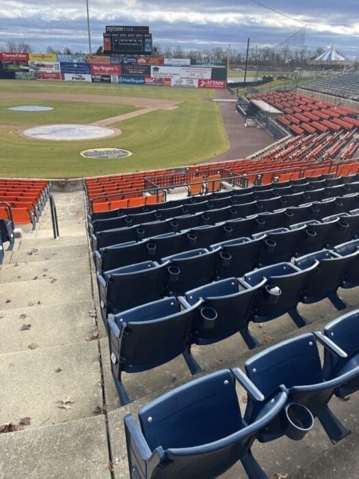 Rows of Empty Navy Blue Stadium Seats with Cup Holders Overlook a Baseball Field Where Fans Eagerly Await the Next Game on the Frederick Keys Schedule the Grass is Patchy with a Scoreboard and Advertising Banners in the Outfield Under Cloudy Skies