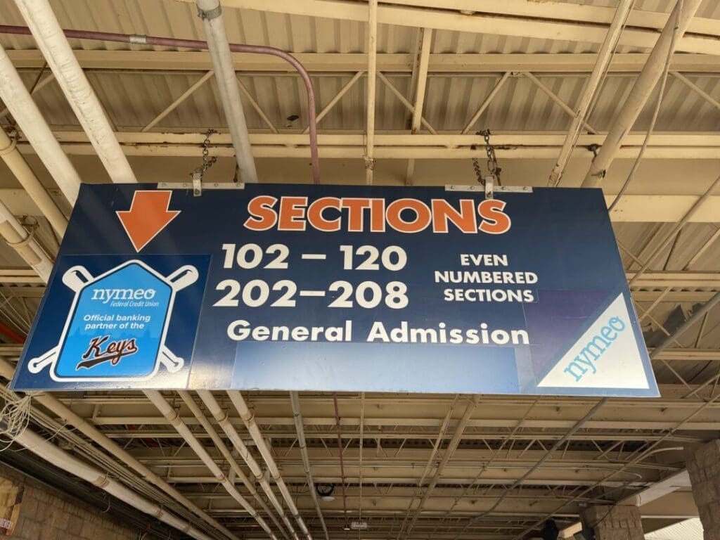 a Large Blue Sign Hanging from the Ceiling Displays Sections with an Orange Arrow Listing Sections 102120 and 202208 Even Numbered and General Admission It Features Nymeo Branding the Keys Baseball Team Logo and Promotes the Frederick Keys Schedule
