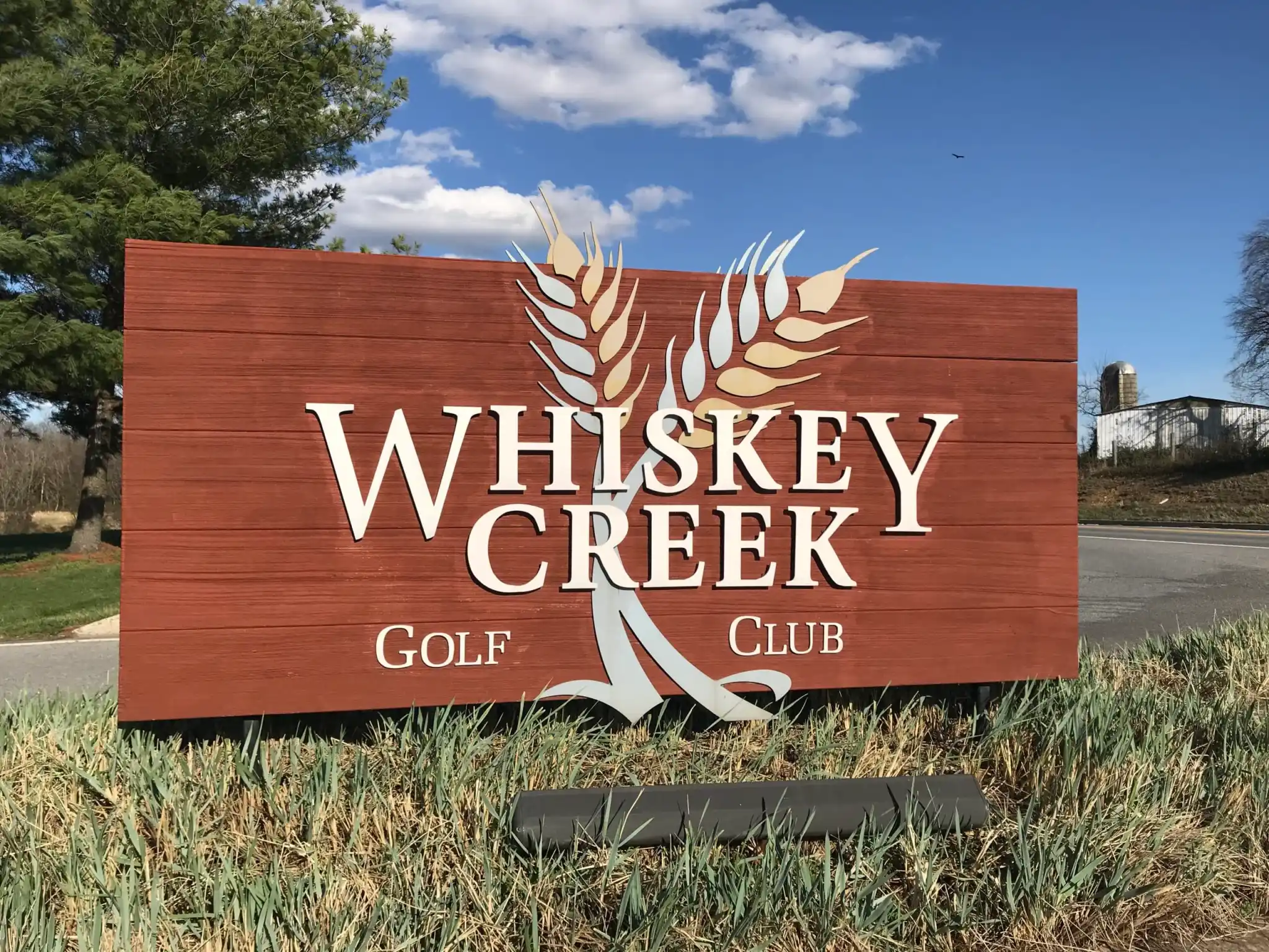 A large wooden sign reading Whiskey Creek Golf Club in white letters, with two stylized wheat stalks crossing behind the text, stands on grass beside a road in New Market MD. Trees, a blue sky with clouds, and distant farm buildings are in the background.