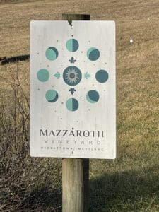 A sign attached to a wooden post reads Mazzaroth Vineyard, Middletown, Maryland—a gem among Frederick MD Wineries. Above the text, phases of the moon circle a sun-like symbol on a weathered white background. Grass and dried bushes frame the scene.
