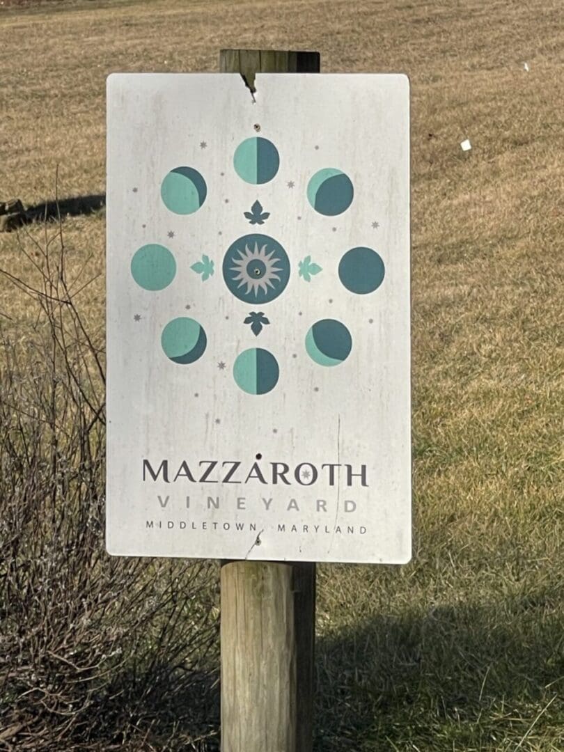 A sign attached to a wooden post reads Mazzaroth Vineyard, Middletown, Maryland—a gem among Frederick MD Wineries. Above the text, phases of the moon circle a sun-like symbol on a weathered white background. Grass and dried bushes frame the scene.
