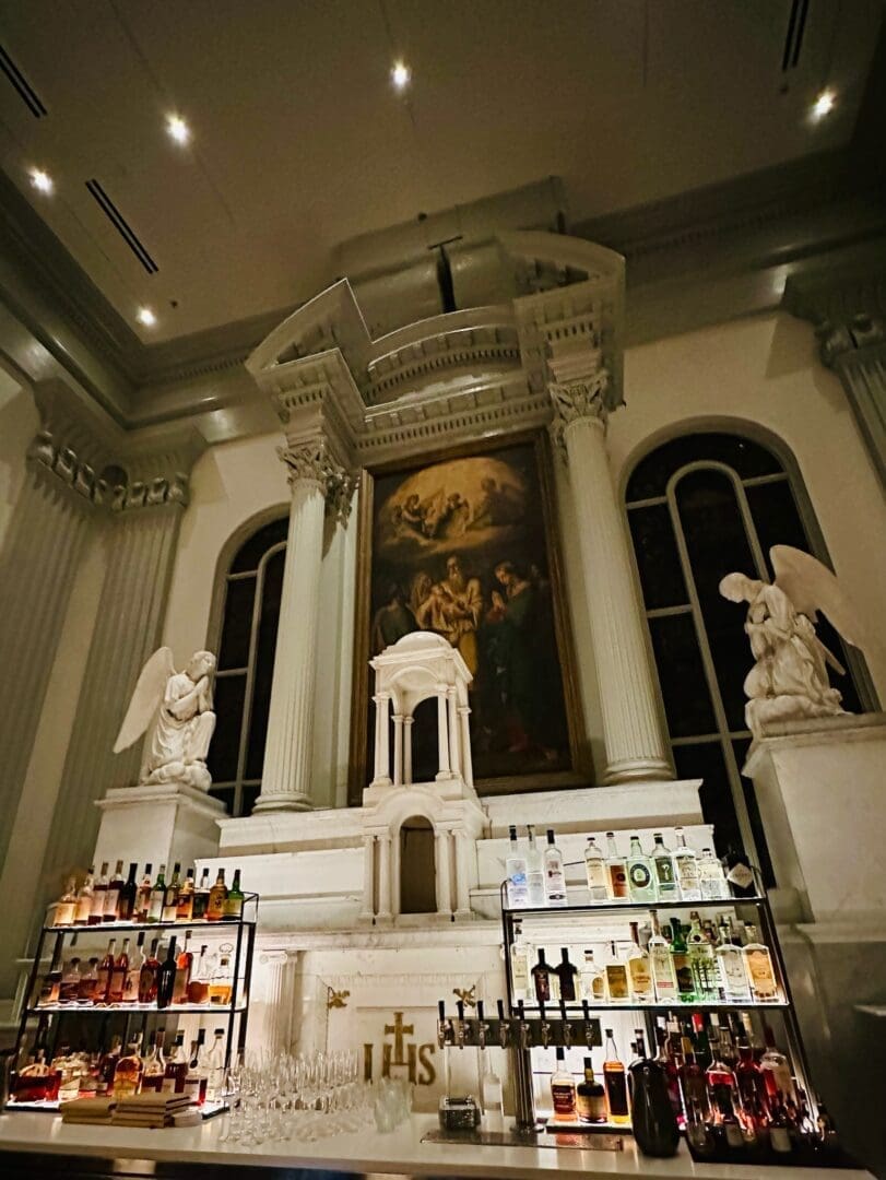 a Grand Bar Reminiscent of the Best Bars in Frederick Stands Before a White Marble Altar with Columns and Angelic Statues Bottles Line Glass Shelves Beneath a Classical Painting While Arched Windows Illuminate the Elegant High Ceilinged Room