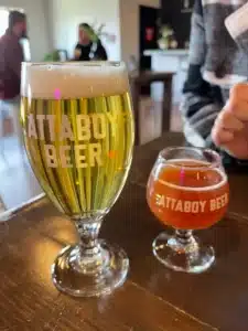 Two glasses of beer on a wooden table feature “ATTABOY BEER” branding, showcasing the vibrant scene of local breweries Frederick offers. A pale golden beer and an amber brew highlight the variety at one of the top breweries in Frederick MD.