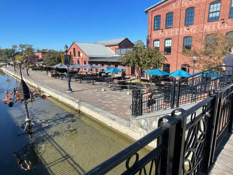 Outdoor dining area with tables and blue umbrellas lines a brick patio next to a calm canal. Steps away, some of the best breweries in Frederick MD offer craft brews, surrounded by red brick buildings, green trees, and a clear blue sky.