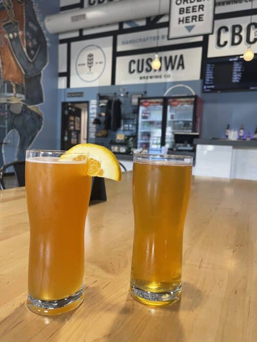 Two tall glasses of beer sit on a light wooden table at one of the best breweries in Frederick MD. One is hazy orange with an orange slice, the other clear golden. Behind them is a taproom with a large menu board and signs reading "CUSTOWA.