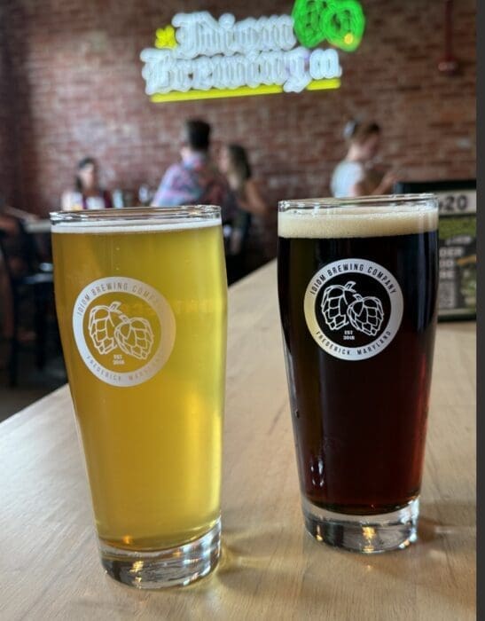 Two pint glasses of beer sit on a wooden table at one of the best breweries in Frederick MD. One beer is pale yellow and hazy, the other dark brown. Both feature a Twin Brewing Company logo, with a blurred brewery sign and people in the background.