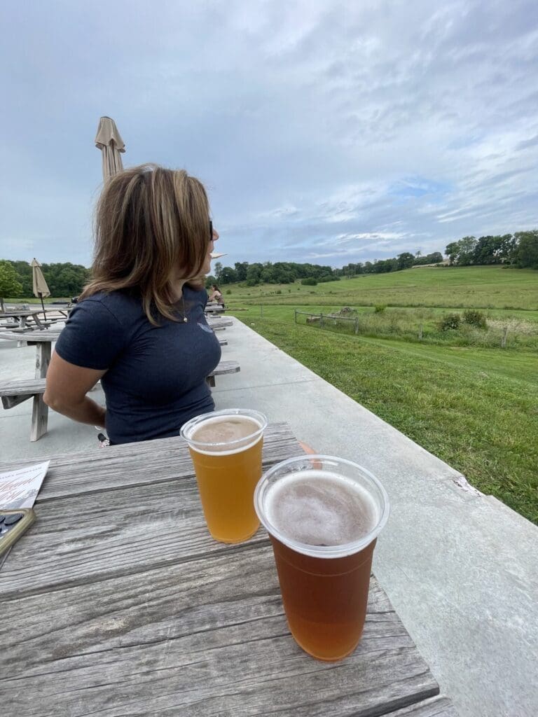 A woman with shoulder-length hair sits at a wooden picnic table outdoors in New Market, MD, gazing toward a green field under a cloudy sky. Two plastic cups of beer rest on the table, with umbrellas and more tables visible in the background.
