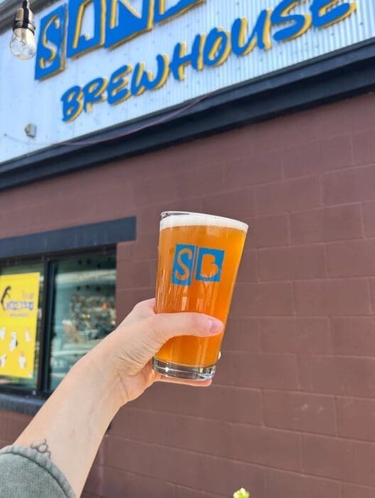 A hand holds a pint of hazy amber beer with a blue SB logo in front of a brown building. A sign above reads SAND BREWHOUSE in bold yellow and blue letters—one of the best breweries in Frederick MD. A tattoo is visible on the person’s wrist.