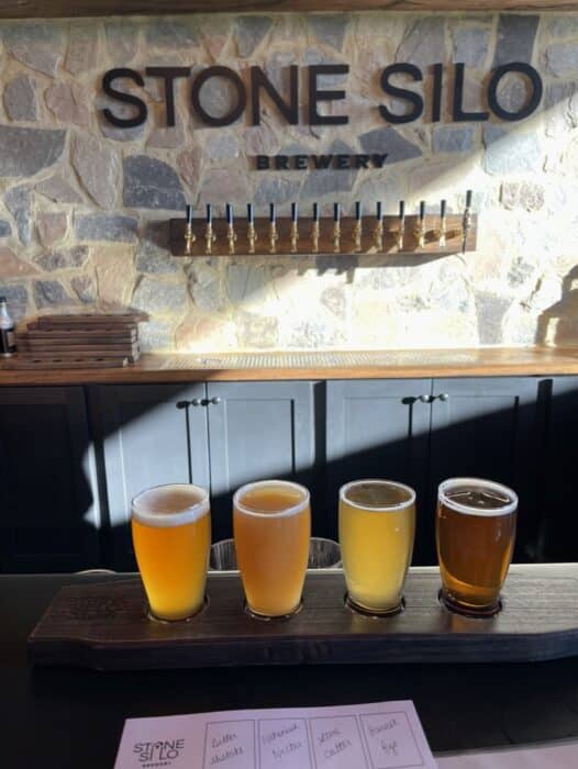 A wooden paddle holds four small glasses of beer in varying shades on a bar counter. Behind them, “STONE SILO BREWERY” is displayed above beer taps, capturing the inviting atmosphere found at the best breweries in Frederick MD. Sunlight casts shadows across the scene.