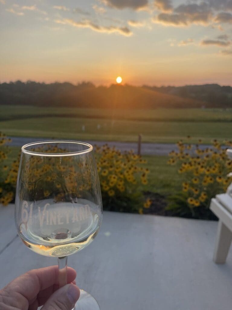 A hand holds a glass of white wine with 6 Vineyard etched on it. Yellow flowers and green fields in New Market, MD, stretch beyond a patio, while the sun sets behind low hills and scattered clouds, creating a warm, tranquil atmosphere.