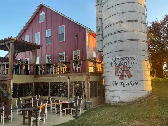 A large red barn with a wooden deck and outdoor seating stands beside a tall white silo labeled “Linganore Winecellars Berrywine” in New Market, MD. The sunny scene and clear sky evoke a peaceful rural vineyard setting.