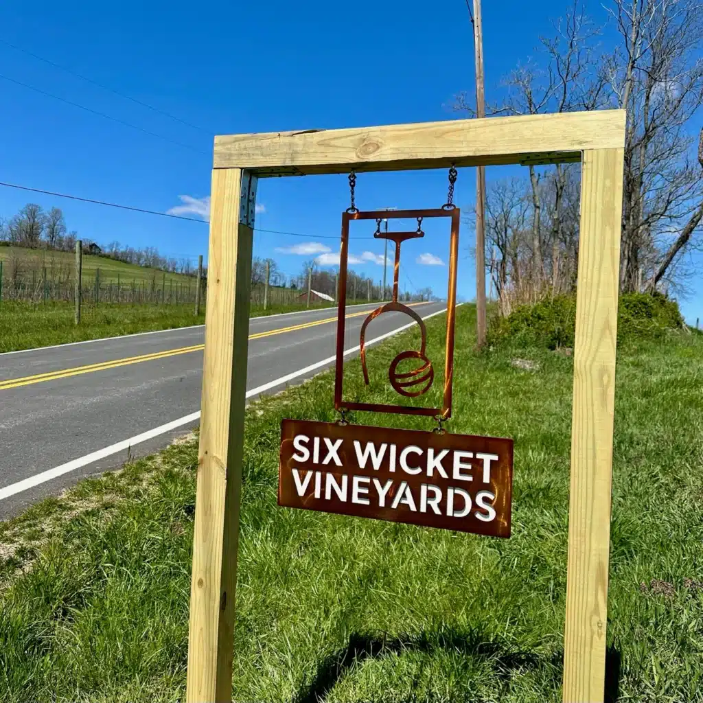 a Wooden Frame Holds a Metal Sign for six Wicket Vineyards by a Rural Roadside Known Among Frederick Md Wineries the Sign Features a Stylized Emblem Above the Name Set on a Grassy Slope with Fences Trees and Blue Sky in the Background