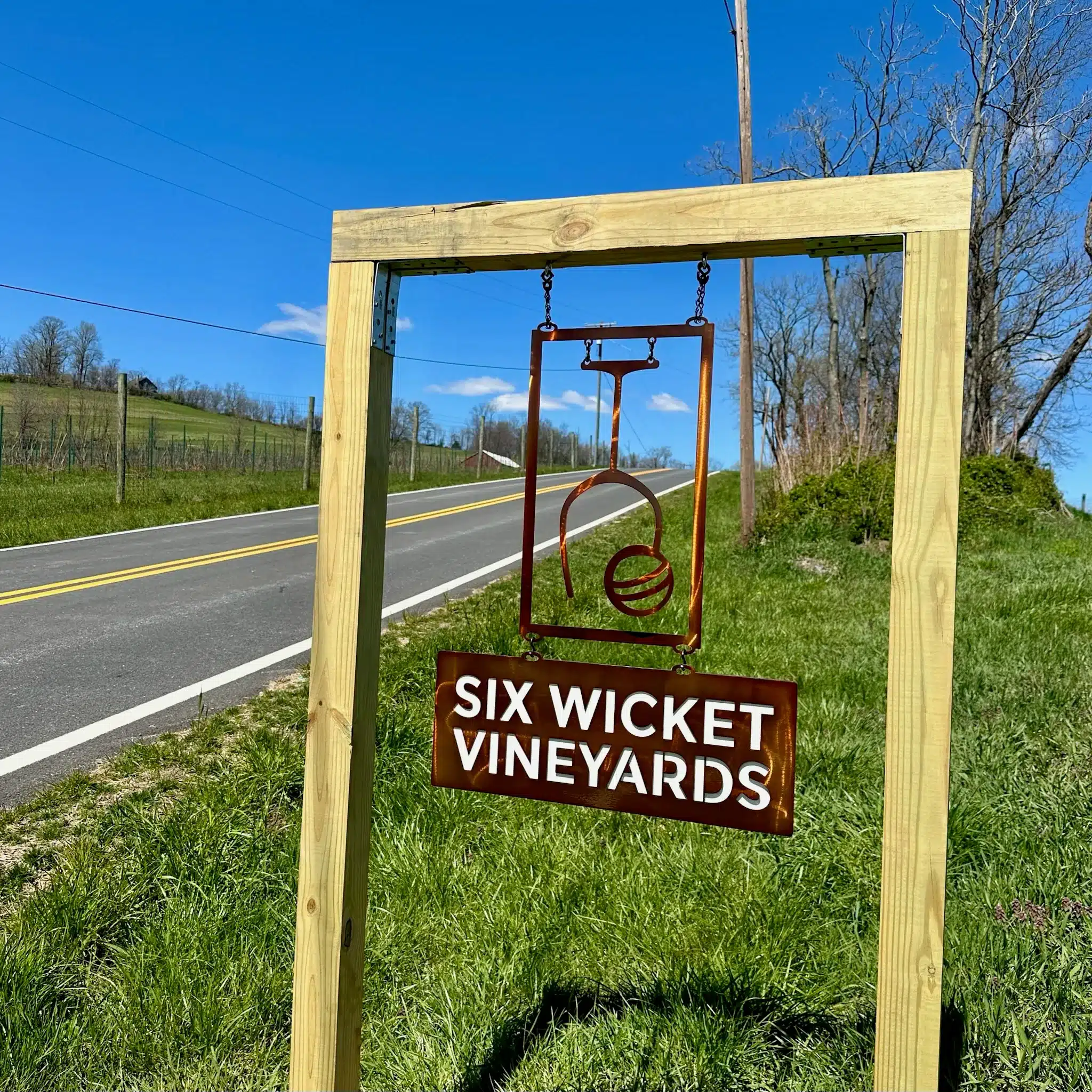 a Wooden Frame Holds a Metal Sign for Six Wicket Vineyards by a Rural Roadside Known Among Frederick Md Wineries the Sign Features a Stylized Emblem Above the Name Set on a Grassy Slope with Fences Trees and Blue Sky in the Background