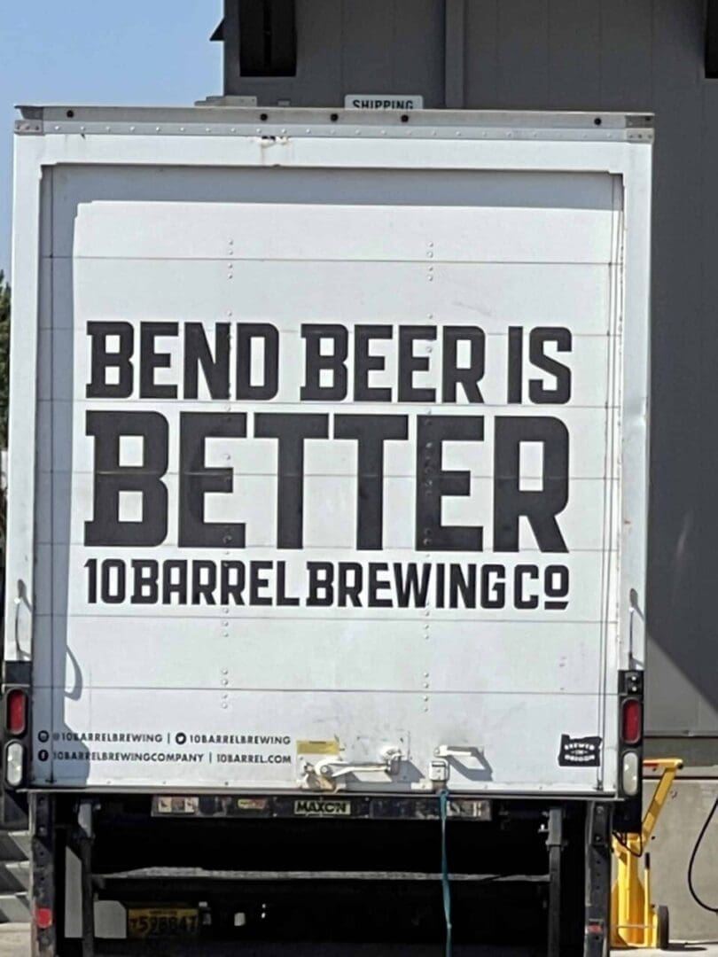 a White Truck with Bold Black Text on the Back Reads Bend Beer is Better 10 Barrel Brewing Co Parked Outdoors Near a Gray Building It Proudly Promotes One of the Top Bend Breweries in Oregon Social Media Handles and a Website Are Displayed Below