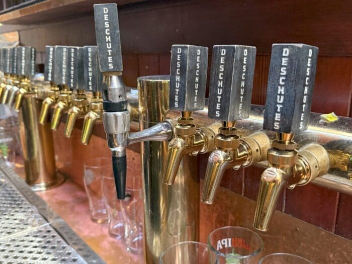 a Row of Brass Beer Taps with Black Handles Labeled deschutes at a Bar Highlights the Rich Craft Scene Among Bend Breweries Several Empty Pint Glasses Sit Below on a Metal Counter with the Taps Mounted on a Wooden Wall Behind