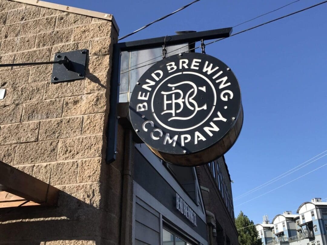 a Circular Black Sign with White Text Reads Bend Brewing Company and Hangs from a Brown Brick Building Showcasing the Logo in the Center This is One of the Renowned Breweries in Bend Oregon Blue Sky and Another Building Are Visible in the Background