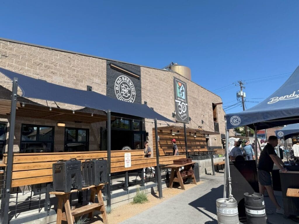 Outdoor Seating Area of One of the Breweries in Bend Oregon with Wooden Benches and Tables Under Black Awnings a Tan Brick Building Displays Brewery Logos and a Water Tank on the Roof As People Enjoy a Sunny Day Beneath a Clear Blue Sky