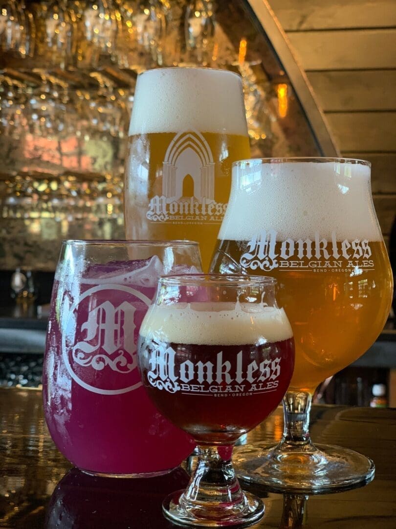Four Glasses of Craft Beer from Monkless Belgian Alesyellow Orange Dark Red and Magentasit on a Reflective Bar Showcasing One of the Top Breweries in Bend Oregon Theyre Set Before a Mirrored Wall Lined with Hanging Glassware