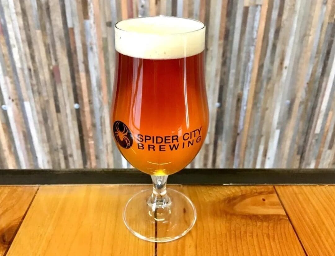 a Tulip Glass Filled with Amber Colored Beer and Topped with a Foamy White Head Sits on a Wooden Surface the Spider City Brewing Logo Graces the Glassa Standout Among Breweries in Bend Oregon Vertical Wooden Planks Add Rustic Warmth to the Background
