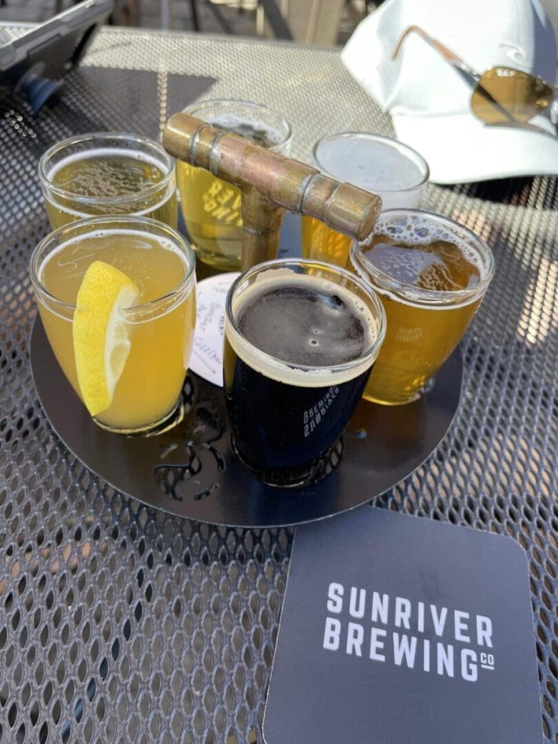 a Round Metal Tray Holds Six Small Glasses of Various Beers from Bend Breweries One Garnished with a Lemon Slice the Tray Sits on an Outdoor Metal Table Next to a Sunriver Brewing Co Coaster with Sunlight and a White Hat in the Background
