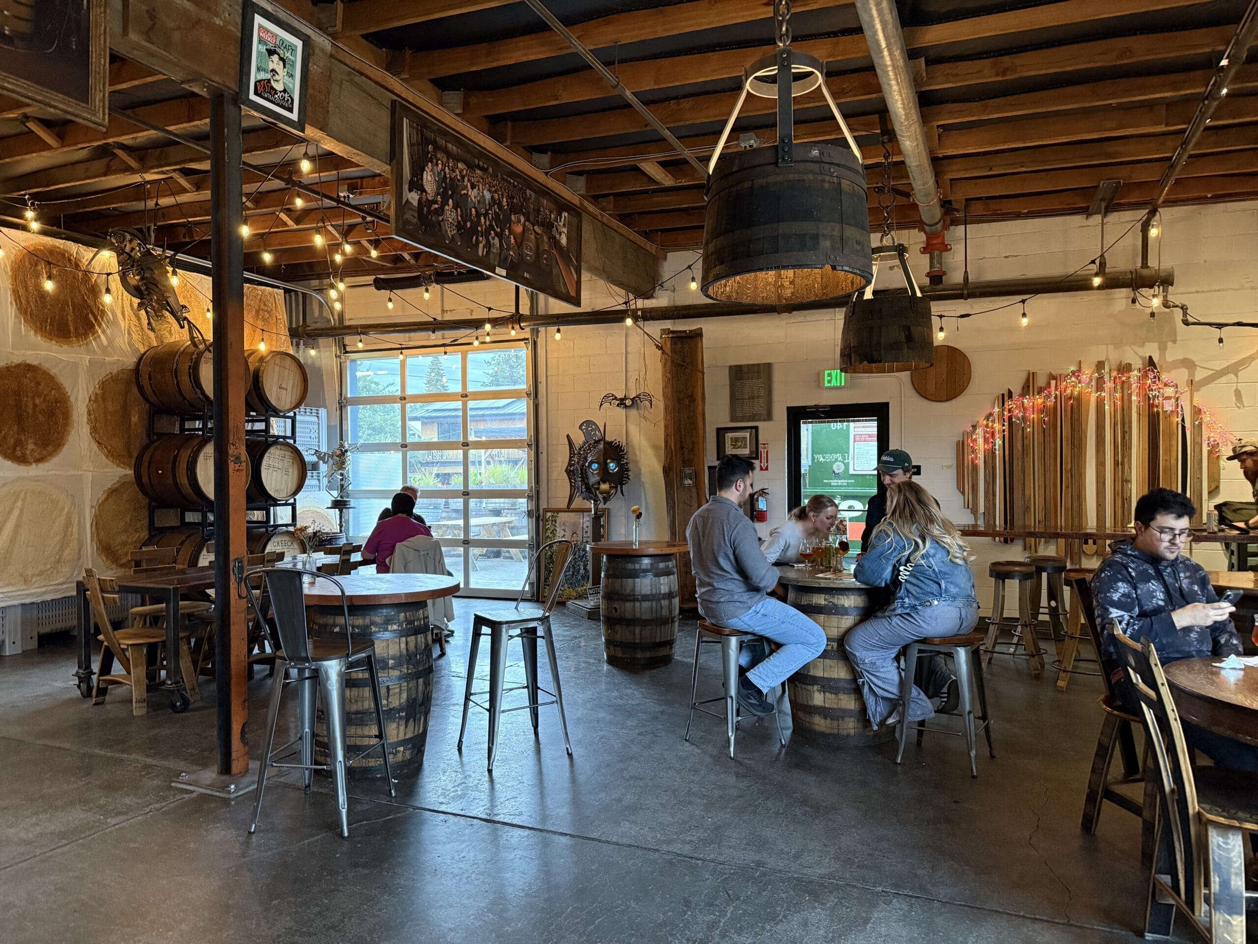 Industrial Style Brewery Taproom with Exposed Beams Hanging Lights and Stacked Barrels on the Left Wall Like Many Breweries in Bend Oregon People Sit at Round Barrel Tables Chatting and Drinking in a Cozy Welcoming Atmosphere Filled with Natural Light