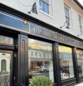 A storefront with black trim and large windows reflects nearby buildings and cars. Above, a gold-trimmed black sign reads UP ON MARKET, one of the best bars in Frederick. The white brick facade features potted plants outside.
