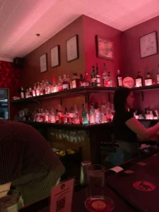 A dimly lit bar with red walls, shelves stocked with bottles of liquor, and framed art—just the kind of cozy spot you’d find in Bend, Oregon. A bartender prepares drinks as a person in a striped shirt sits at the bar in the foreground.