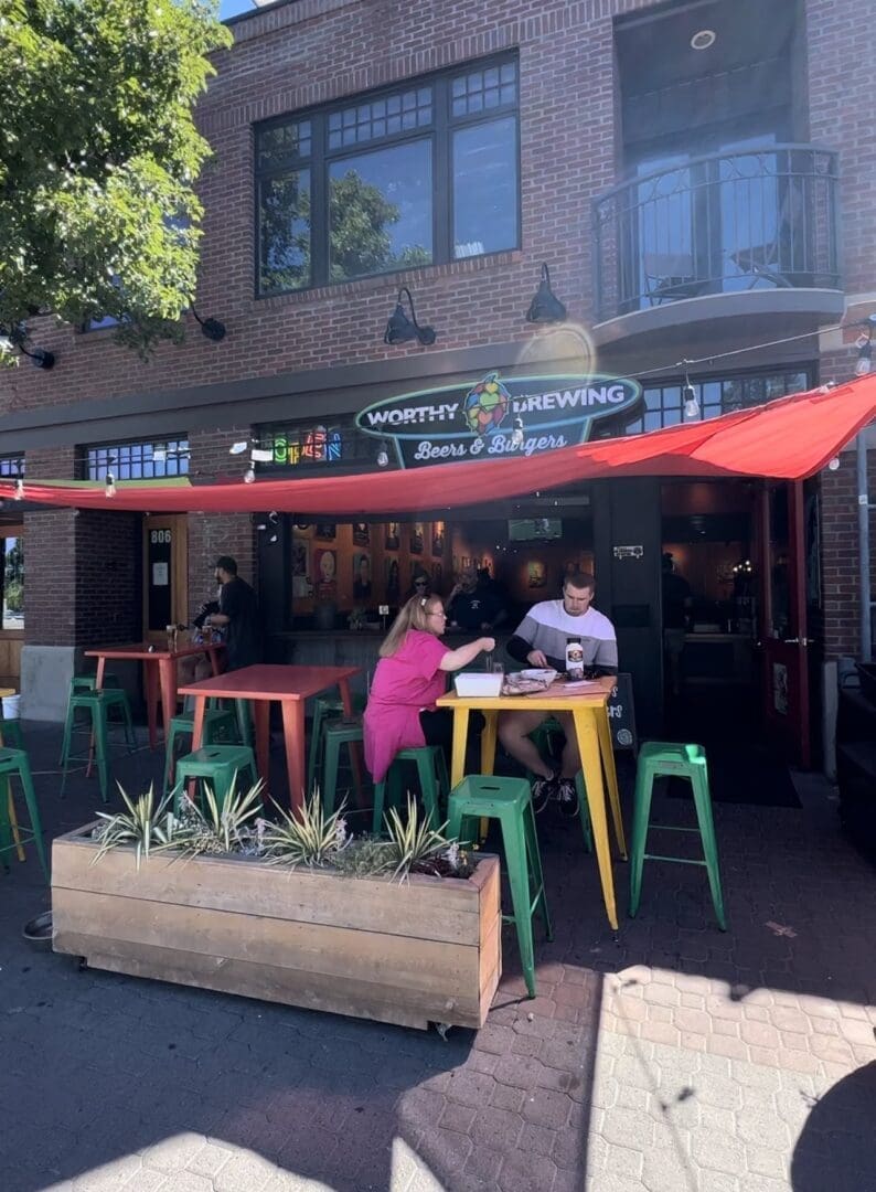 Two People Sit at a Colorful Outdoor Table Under a Red Canopy in Front of Worthy Brewing One of the Top Breweries in Bend Oregon the Man Wears a Striped Shirt the Woman Wears Bright Pink Green Stools and Spiky Plants Add Charm As Sunlight Casts Shadows