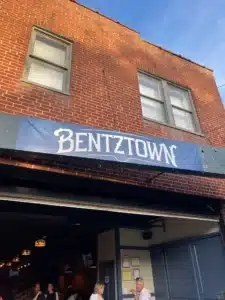 A brick building with two windows displays a blue sign reading BENTZTOWN in white, stylized letters. Below, people are seated at tables inside the warm-lit space—one of the best bars in Frederick—visible through a large open window.
