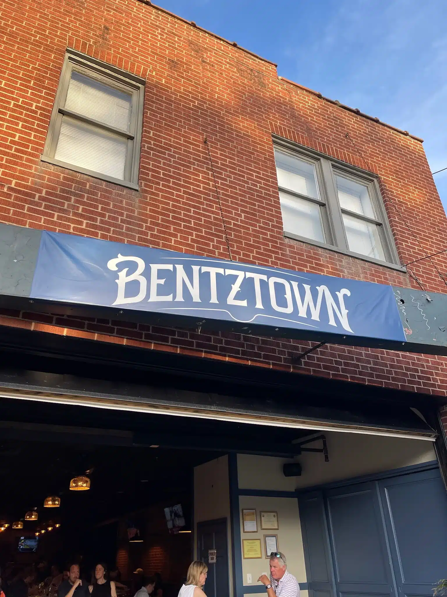 a Brick Building with Two Windows Displays a Blue Sign Reading Bentztown in White Stylized Letters Below People Are Seated at Tables Inside the Warm lit Spaceone of the Best Bars in Frederickvisible Through a Large Open Window