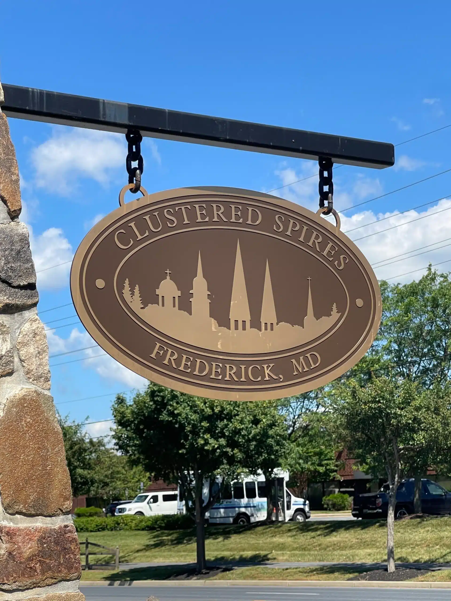 Oval tan sign reading “CLUSTERED SPIRES FREDERICK, MD,” featuring church spires silhouettes, hangs from a metal bracket on a stone pillar. In the background are trees, parked white vans, and a blue sky with clouds near New Market, MD.