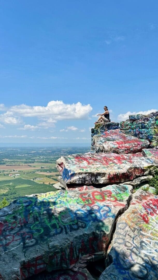 A person sits on the edge of a layered, rocky cliff covered in colorful graffiti—one of the best hikes around Frederick MD—overlooking a wide expanse of farmland and green fields under a bright blue sky with scattered clouds.