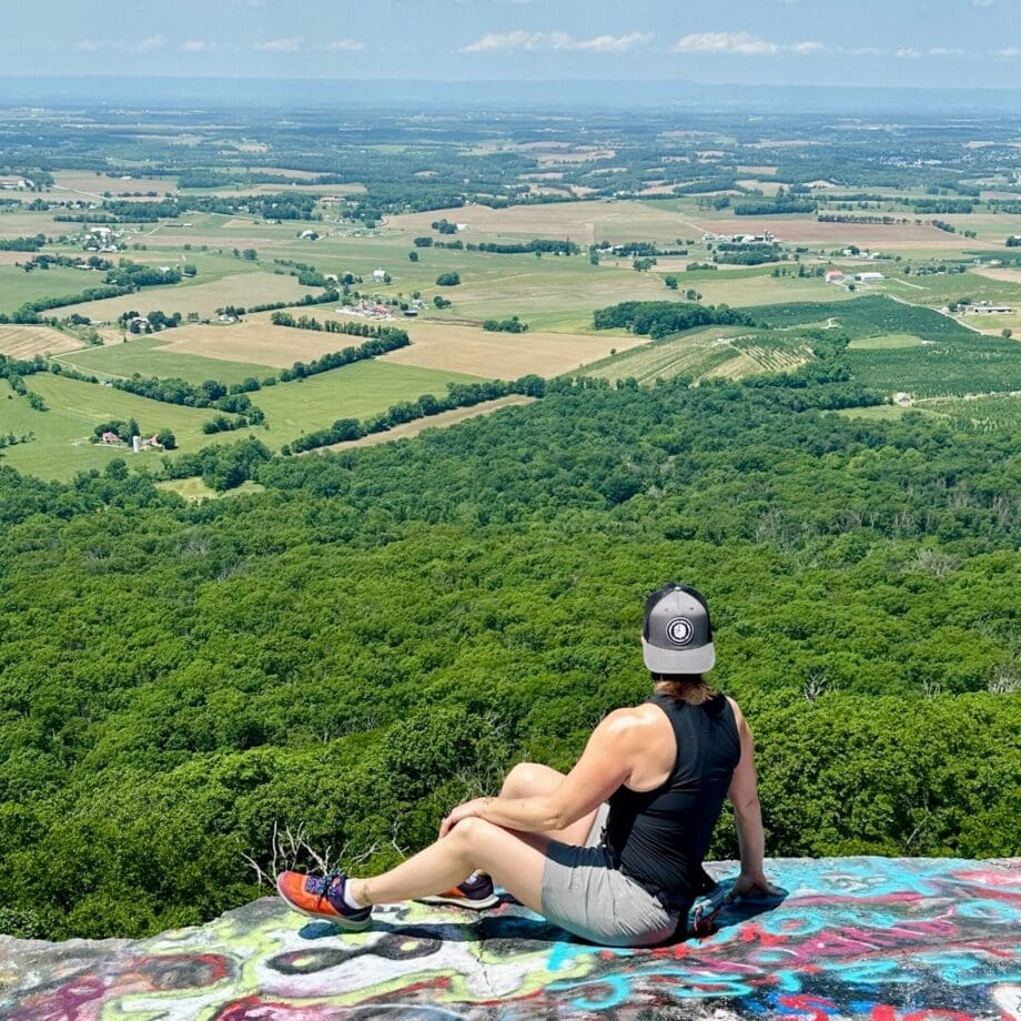 A person in a black tank top, gray shorts, and a cap sits on a colorful graffiti-covered rock—one of the best hikes around Frederick MD—taking in sweeping views of farmland, fields, distant hills, and lush green forest under a clear sky.